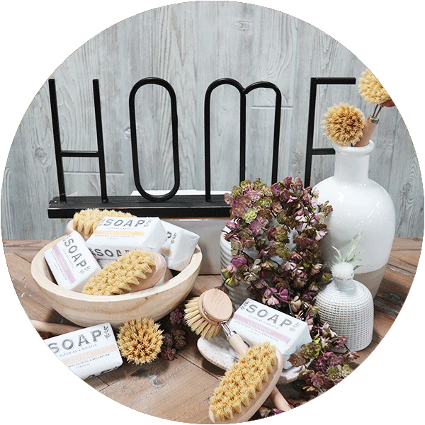 A collection of soap bars, scrub brushes and artificial foliage on a wooden table with a gray wood background.